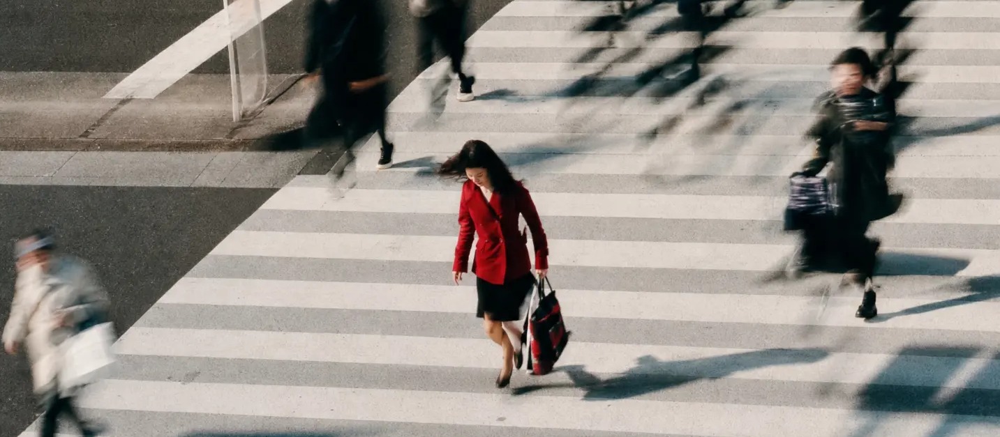 A woman in a red blazer walking across a busy crosswalk seen from above, the crowd around her blurred in motion